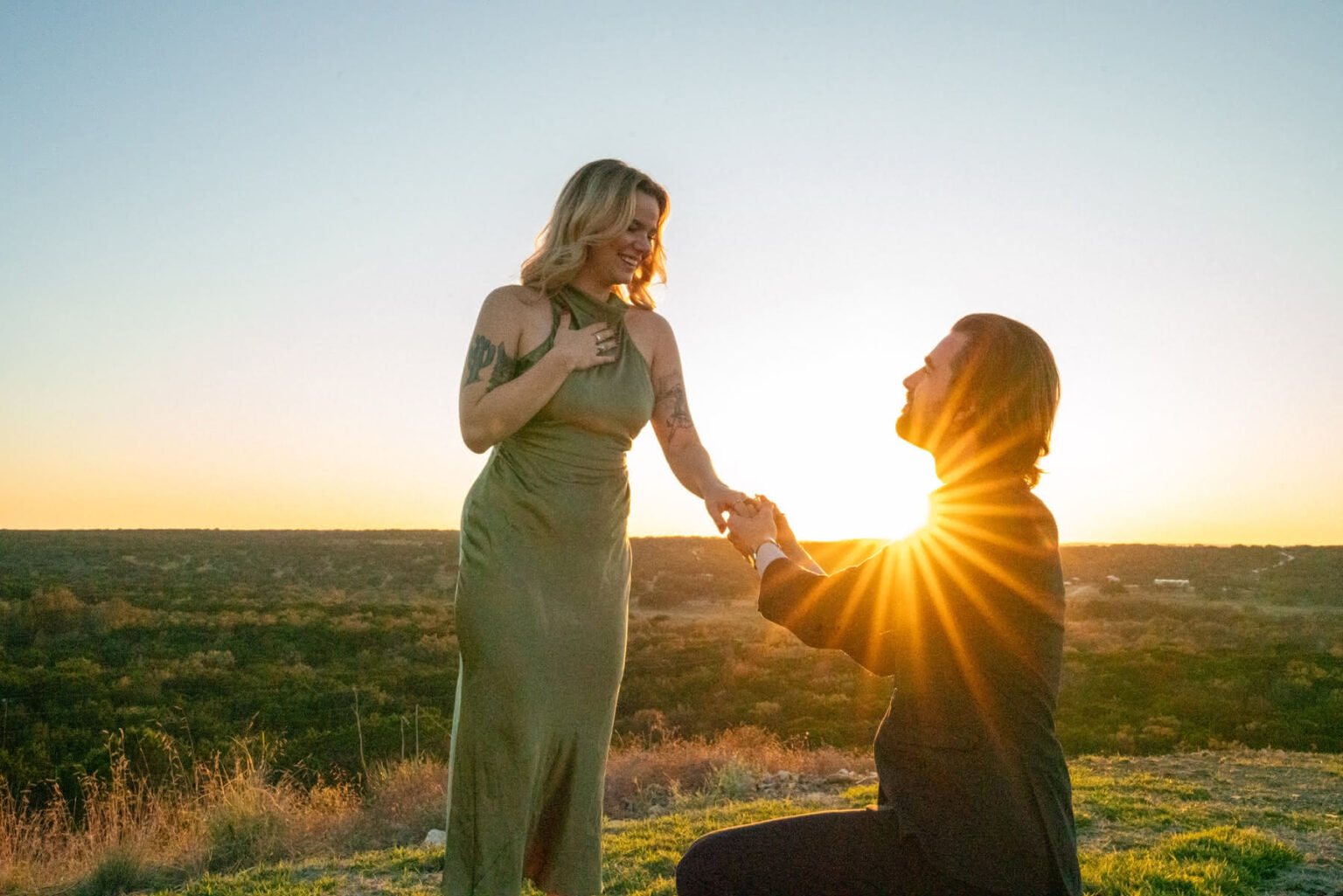 Man in a suit gets down on one knee to propose to his girlfriend on Hill Country Vista at sunset